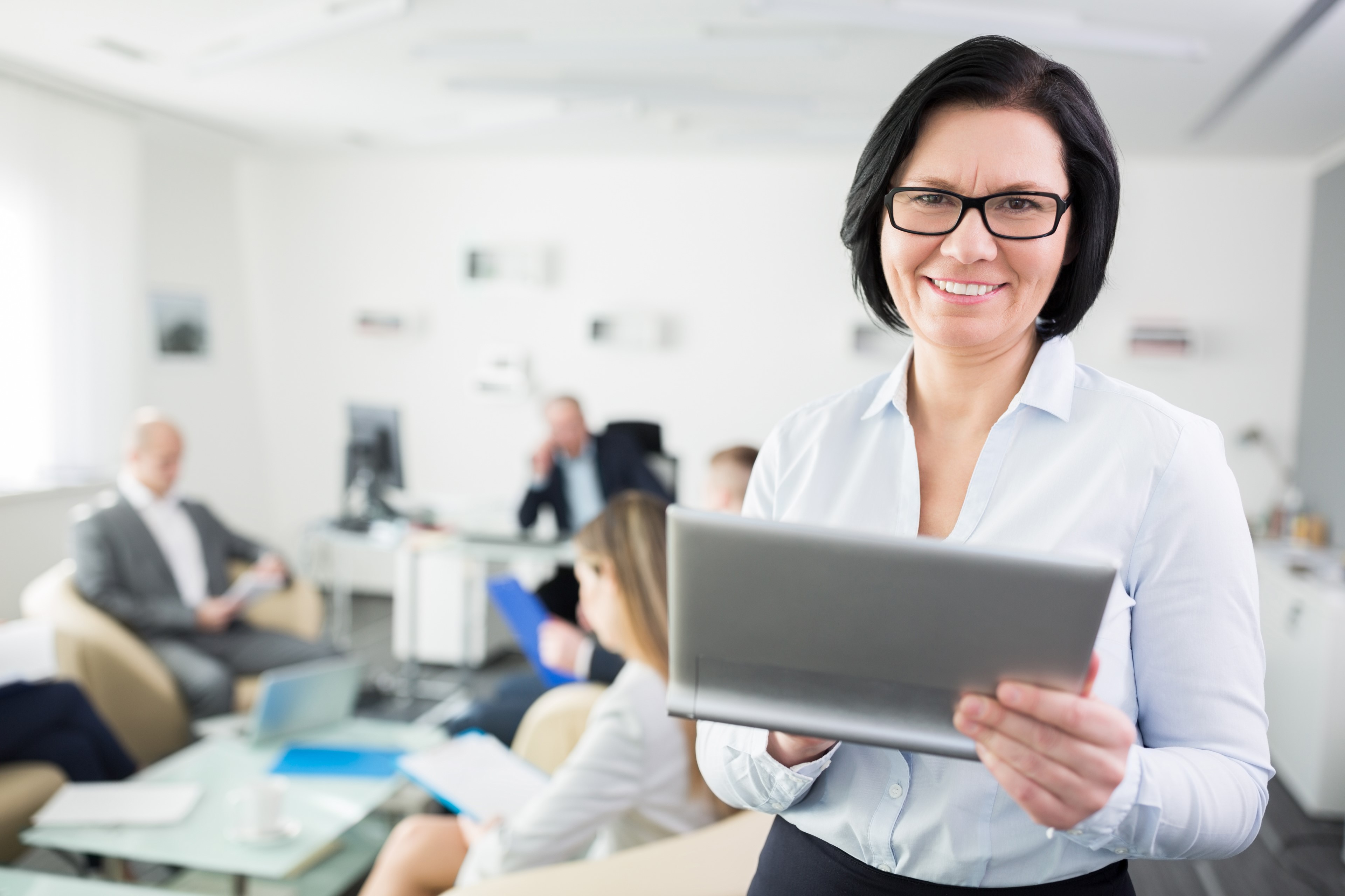 Close up photo of a middle aged business woman holding a tablet. Background is blurry with a multigenerational team sitting down around low tables with documents and computers in front of them.